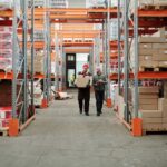 Two workers handle a package in a spacious warehouse surrounded by shelves stocked with boxes and products.